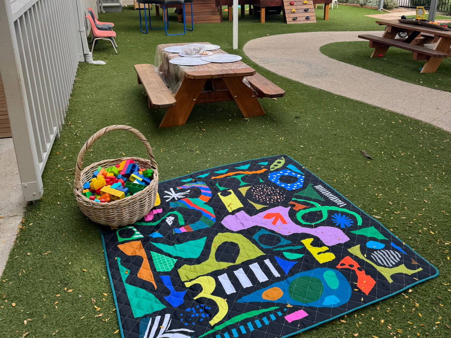 Colorful rug with geometric patterns on grass, picnic table, and outdoor furniture in the background of a childcare centre