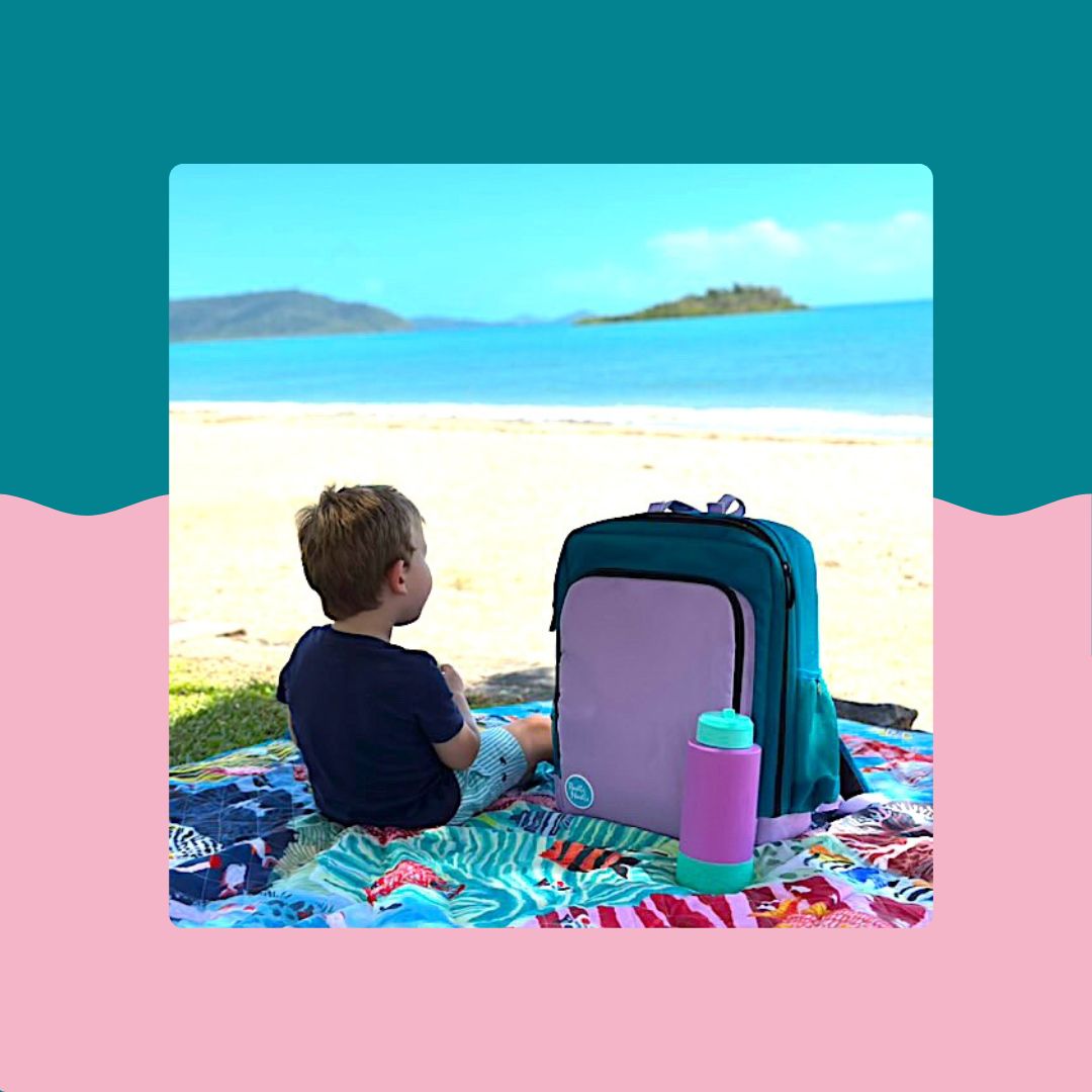 toddler on colourful rudie nudie picnic rug at the beach beside the teal and lilac parents nappy backpack