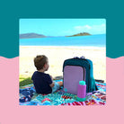 toddler on colourful rudie nudie picnic rug at the beach beside the teal and lilac parents nappy backpack