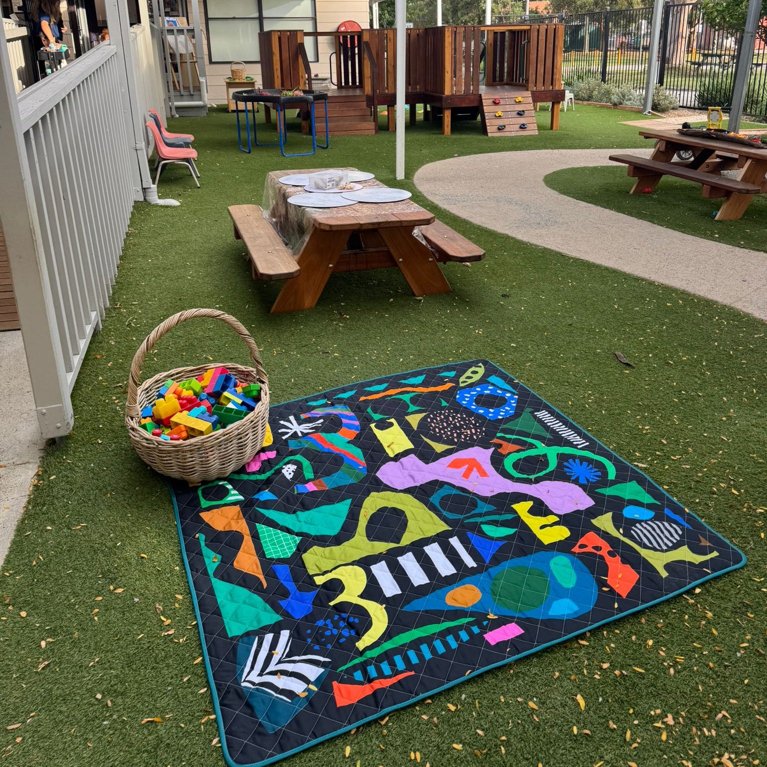 Colorful rug with geometric patterns on grass, picnic table, and outdoor furniture in the background of a childcare centre